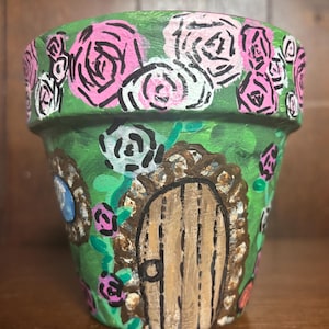 May include: Hand-painted terracotta flower pot featuring a whimsical fairy house design. The pot is painted green with pink and white roses around the rim and a wooden door with a stone frame. A charming addition to any garden.