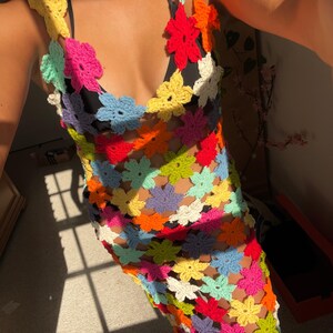 May include: A colorful crochet dress featuring a floral pattern in various bright colors. The dress has a halter neckline and a fitted silhouette. The crochet work is open, creating a see-through effect. The dress is perfect for a summer day.