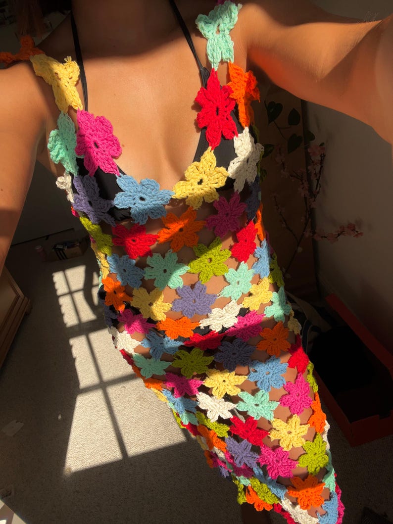 May include: A colorful crochet dress featuring a floral pattern in various bright colors. The dress has a halter neckline and a fitted silhouette. The crochet work is open, creating a see-through effect. The dress is perfect for a summer day.