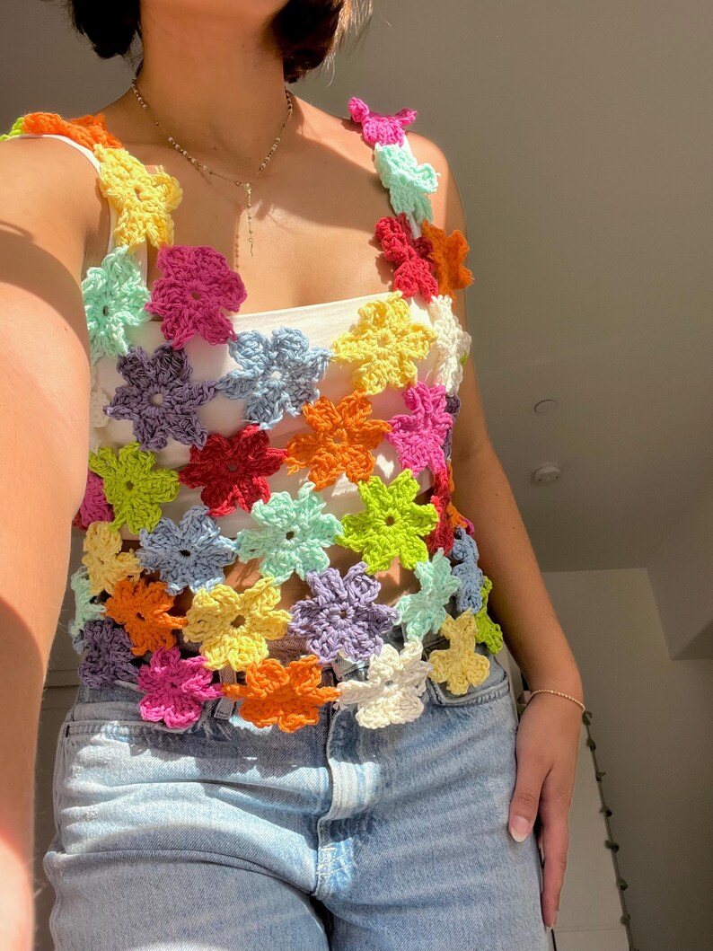 May include: A colourful crochet top with a floral design. The top is made of a variety of bright colours, including pink, yellow, green, blue, and orange. The top is sleeveless and has a scoop neck. The top is worn over a white vest top.