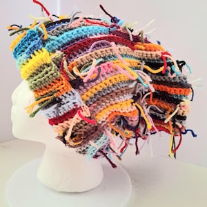 May include: A multi-colored crochet beanie featuring a textured, striped pattern in shades of yellow, orange, blue, red, purple, and brown. The hat is adorned with numerous loose yarn ends and frayed threads throughout, creating a shaggy, layered appearance.