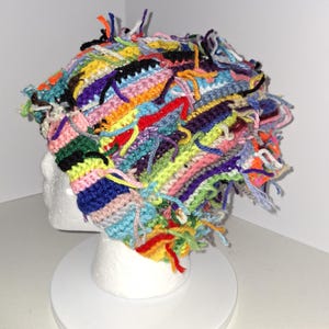 May include: A multi-colored crochet beanie featuring a textured, shaggy design with numerous loose yarn ends in shades of neon green, bright blue, red, yellow, purple, pink, and black, constructed with varied stitch patterns throughout the fabric.