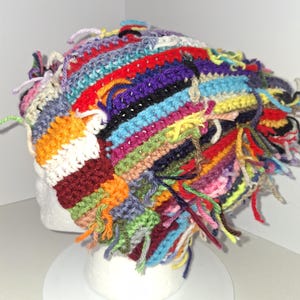 May include: A multi-colored crochet beanie featuring horizontal stripes in red, blue, yellow, green, orange, purple, and white. The hat is constructed with visible yarn ends and loose threads throughout, creating a textured, shaggy appearance.