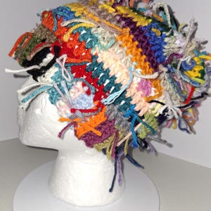 May include: A multi-colored crochet beanie featuring a patchwork design with various yarn textures and loose fringe ends. The hat is constructed from a vibrant mix of yellow, orange, red, blue, green, purple, and white yarn in diverse stitch patterns.