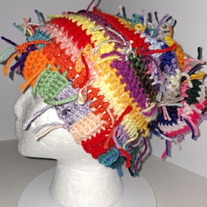 May include: A multi-colored crochet beanie featuring a vibrant array of yarn colors including red, yellow, orange, purple, and green. The hat is constructed with a textured stitch pattern and is adorned with numerous loose yarn ends throughout.