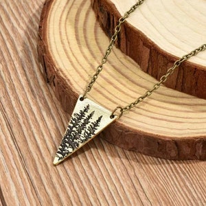 May include: A gold-toned necklace with a triangular pendant featuring a silhouette of a forest. The pendant is made of metal and has a rustic, vintage look.