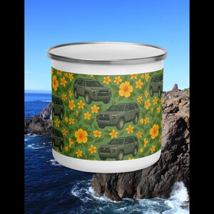 Könnte beinhalten: Weißer Emaillebecher mit grünem Hintergrund, der olivgrüne SUVs und gelbe Blumen zeigt. Der Becher hat einen silbernen Rand und eine weiße Innenseite. Das Design umschließt den gesamten Becher und erzeugt ein zusammenhängendes Muster.