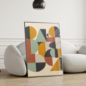 Puede incluir: Impresión artística enmarcada con un diseño abstracto de formas geométricas en naranja, amarillo, verde y negro. La obra de arte incluye círculos, triángulos y semicírculos sobre un fondo crema. La impresión se muestra en un suelo de madera, apoyada en un sofá blanco.