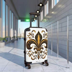 May include: White suitcase with a black and gold fleur-de-lis design. The suitcase has a black handle and four wheels. The text "Ouest & Plus" is printed on the suitcase.