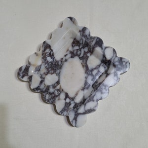 May include: A white and black marbled square dish with a scalloped edge. The dish is made of natural stone and has a unique, organic shape.