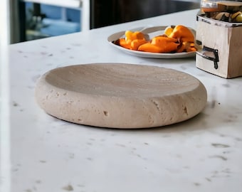 May include: A light brown, round, stone serving dish with a textured surface. The dish is on a white countertop.