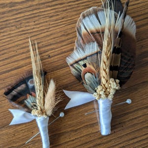 May include: Two boutonnieres featuring brown and tan pheasant feathers, dried wheat stalks, and small clusters of dried cream-colored flowers. Each stem is wrapped in white satin ribbon with a white bow and includes a silver pin with a pearl head.