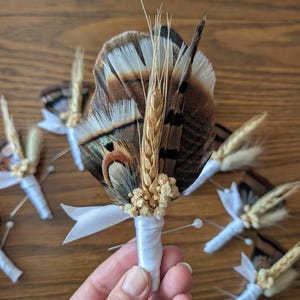 May include: A boutonniere featuring brown and white patterned feathers, a stalk of dried wheat, and small clusters of dried cream-colored flowers. The stems are wrapped in white satin ribbon with a small bow and a silver pin attached to the base.
