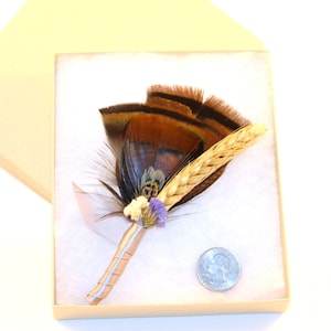 May include: A brown and black pheasant feather boutonniere with a wheat stalk and small purple and white flowers. The boutonniere is wrapped in a light brown ribbon and is displayed in a small box.