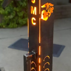 May include: A metal fire pit with a cutout design of the USMC logo and the letters "USMC" on the side. The fire pit is lit and has a bright orange flame.