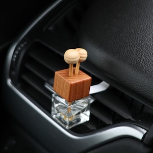 May include: A car air freshener with a wooden block and two wooden ball accents. The air freshener sits on a car vent, with a clear glass bottle base. The car interior is black and silver.