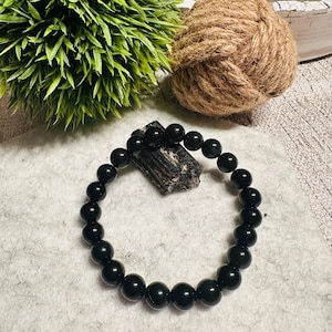 Puede incluir: Un brazalete negro hecho de cuentas redondas y lisas. El brazalete est&aacute; sobre una superficie gris claro.