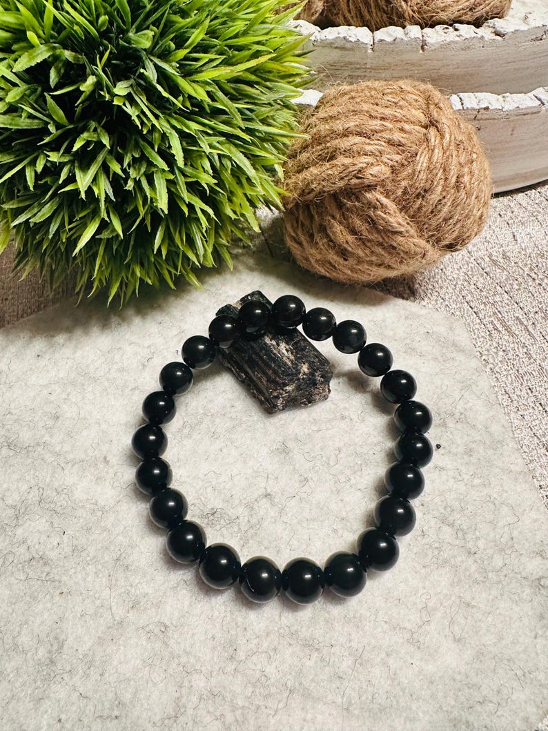 Puede incluir: Un brazalete negro hecho de cuentas redondas y lisas. El brazalete est&aacute; sobre una superficie gris claro.