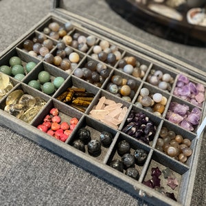 May include: A gray fabric-lined box with compartments filled with various types of crystals and stones, including amethyst, rose quartz, agate, and tiger's eye.