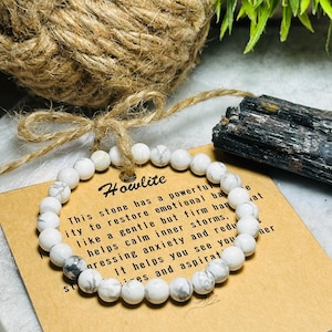 May include: White howlite beaded bracelet on a brown card with the text "Howlite" and a description of the stone's properties. This stone has a powerful ability to restore emotional balance, like a gentle but firm hand. It helps calm inner storms, pressing anxiety and reducing stress. It helps you see your true self and aspirations.
