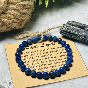 May include: A blue lapis lazuli beaded bracelet with a brown card that reads "Lapis Lazuli" and describes the stone's properties. The card is sitting on a white surface.