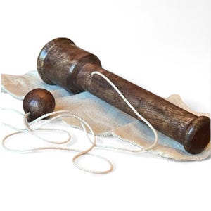 May include: A wooden ball and cup toy, also known as a bilboquet, rests on a beige cloth. The toy features a dark brown, cylindrical handle with a ball attached by a white string. The ball is also dark brown.