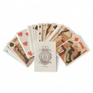 May include: A deck of playing cards fanned out on a white surface. The cards feature red, black, and brown designs, including hearts, spades, clubs, and diamonds. A small card in the center displays text and a crest.
