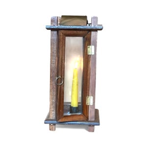 May include: A wooden lantern with a lit yellow candle inside. The lantern has a rectangular shape with a hinged door and a metal ring handle. The frame is made of dark wood, and the glass panels allow the candle's light to shine through.