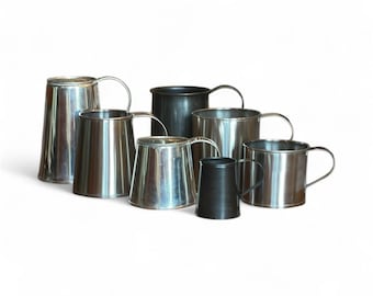May include: A selection of metal mugs in different sizes and finishes. Some are polished silver, others are matte black. Each mug features a simple, curved handle. The mugs are displayed against a white backdrop.