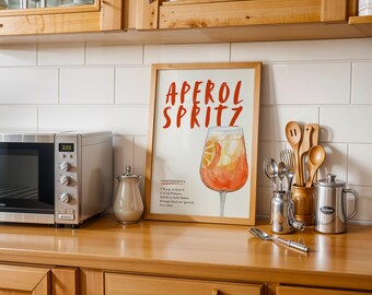 Pode incluir: Uma impressão emoldurada com uma ilustração em aquarela de um coquetel Aperol Spritz. A impressão tem o texto "Aperol Spritz" em letras vermelhas e lista os ingredientes do coquetel.