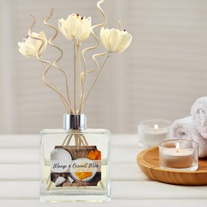 May include: A clear glass diffuser bottle with a silver cap, filled with a yellow liquid. The label reads "Mango & Coconut Milk." Decorative flower reeds and a wooden tray with candles and a rolled towel are in the background.
