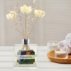 May include: A clear glass reed diffuser with a silver collar, featuring decorative cream-colored flower-shaped reeds. The label reads "Emerald Isle." The diffuser is displayed with candles and a wooden tray.