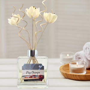 May include: A clear glass Nag Champa reed diffuser with decorative flower stems. The diffuser has a silver collar and a label that reads "Nag Champa". The image includes a lit candle and a wooden tray.