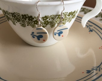 May include: A pair of round, ceramic earrings with a white base and a blue and red floral design. The earrings hang from silver-colored hooks. The earrings are displayed in front of a white teacup and saucer with a green floral pattern.