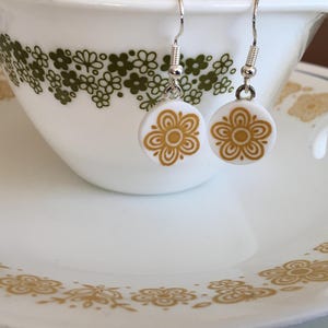 May include: A pair of white ceramic earrings with a gold floral design. The earrings are hanging from silver hooks.