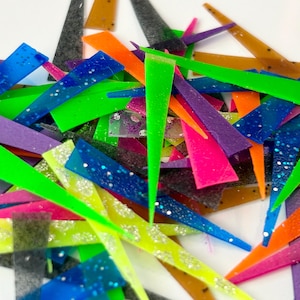 May include: A pile of colorful, glittery, triangular-shaped plastic pieces. The pieces are in various shades of green, blue, purple, pink, orange, and yellow. Some pieces are translucent, while others are opaque.
