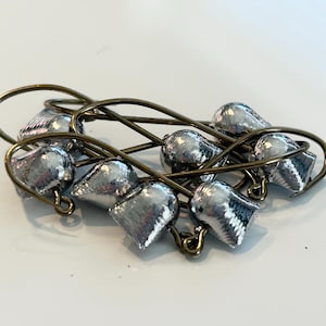 May include: A collection of silver-colored fishing jig heads with bronze-colored wire hooks. The jig heads have a teardrop shape and are clustered together. The hooks are curved and appear to be made of a sturdy metal.