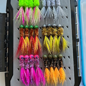 May include: A black tackle box with a gray foam insert containing 12 colorful fishing lures. The lures have different colored feathers, beads, and bodies.