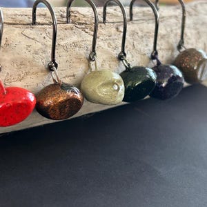 Könnte beinhalten: Sechs Angel-Jigköpfe in verschiedenen Farben, darunter Rot, Kupfer, Olive, Dunkelgrün, Lila und Braun. Jeder Jigkopf ist an einem schwarzen Haken befestigt. Die Jigköpfe werden auf einem Stück Treibholz präsentiert.