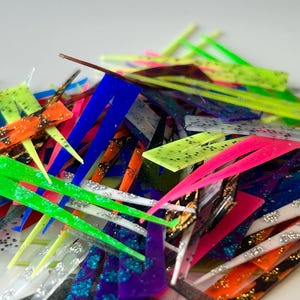 May include: A pile of colorful, glittery plastic lure blades in various shapes and sizes. The blades are a mix of bright colors, including pink, green, blue, yellow, and orange.