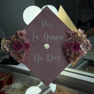 Op de afbeelding: Een afstudeerhoed in een stoffige roze kleur, versierd met bloemstukken in roze en paarse tinten, en kleine vlinders. De hoed heeft de tekst "Por La Gracia De Dios" in zilveren letters.