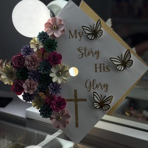 Op de afbeelding: Een versierde afstudeerhoed met de woorden "My Story His Glory". De hoed heeft papieren bloemen in verschillende kleuren, gouden glittervlinders en een gouden glitterkruis. De bloemen zijn roze, paars, wit en groen.