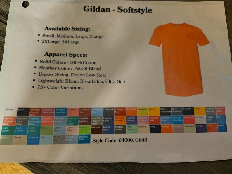 以下が含まれることがあります： オレンジ色のGildan Softstyle Tシャツが白い背景に表示されています。画像には、利用可能なサイズ、アパレル仕様、75色以上のカラーバリエーションの詳細が記載されたテキストが含まれています。スタイルコード：64000、G640。