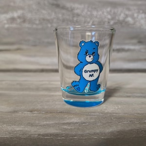 May include: Clear shot glass featuring a blue cartoon bear with a grumpy expression and the text "Grumpy AF". The glass has a blue base and is set against a grey wooden backdrop. The shot glass is a common item.