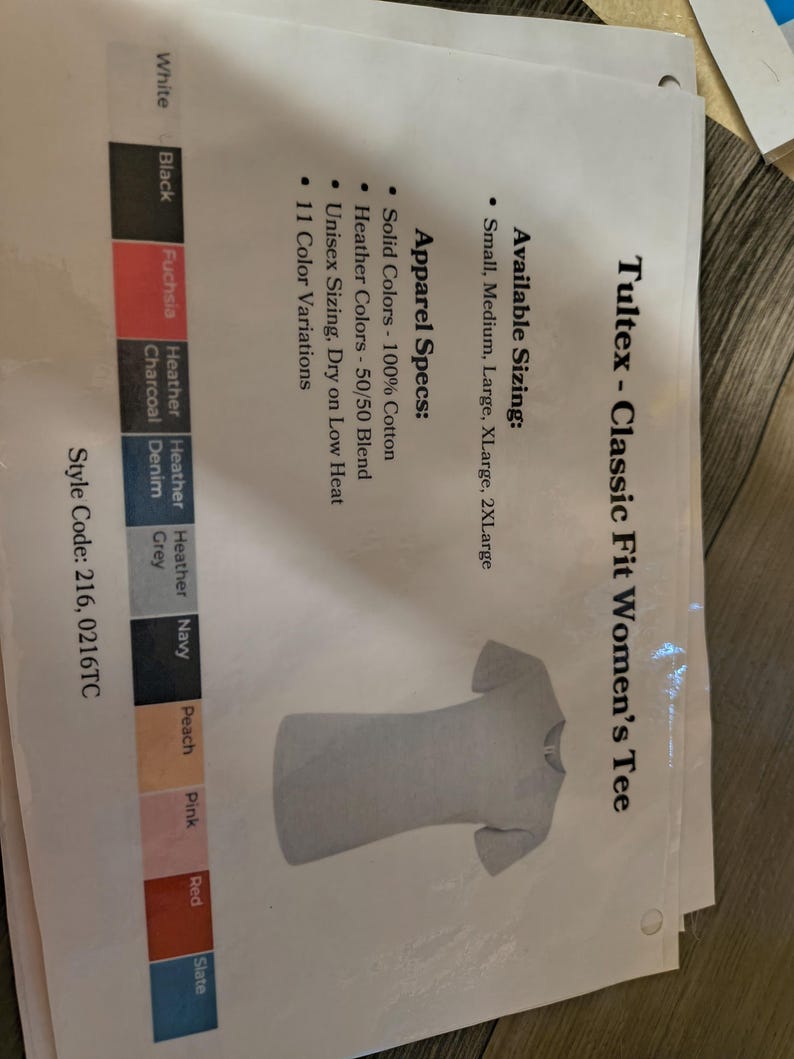 以下が含まれることがあります： 白紙に表示された、ライトグレーのTultex Classic Fit Women's Tee。利用可能なサイズ、アパレル仕様、色のバリエーションも記載されています。このシャツは半袖、クルーネックです。