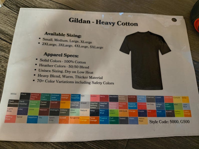 以下が含まれることがあります： 黒いTシャツのグラフィック、カラーサンプル、テキストが印刷されたシート。テキストには「Gildan - Heavy Cotton」、サイズオプション、アパレル仕様、スタイルコードが含まれています。カラーオプションには、白、黒、さまざまな色合いが含まれています。