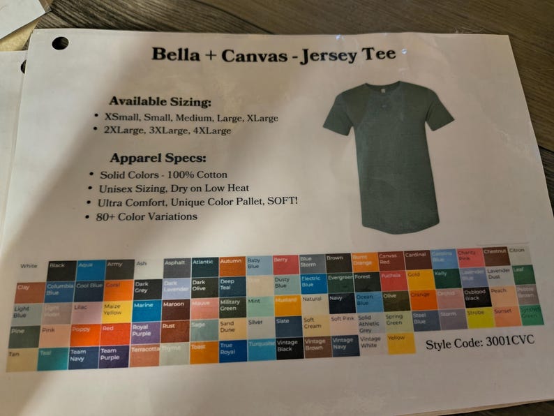 以下が含まれることがあります： Bella + Canvas ジャージーTシャツの製品シート。シートには、サイズオプション、アパレル仕様、80以上のバリエーションのカラーパレットが含まれています。右側にはグリーンのTシャツが表示されています。スタイルコード3001CVC。
