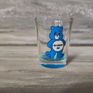 May include: A clear shot glass with a blue base and a blue cartoon bear graphic. The bear has a grumpy expression and the text "Grumpy AF" is printed on the bear's belly. The shot glass is on a gray wooden surface.