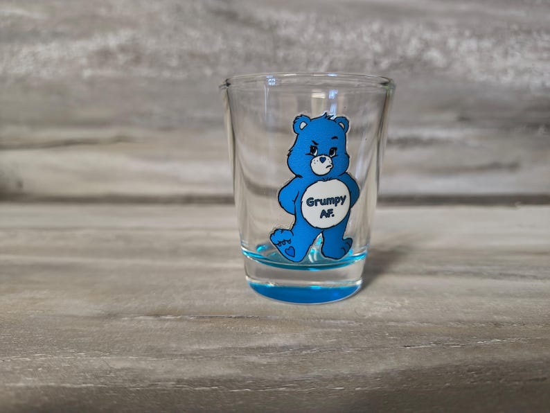 May include: A clear shot glass with a blue base and a blue cartoon bear graphic. The bear has a grumpy expression and the text "Grumpy AF" is printed on the bear's belly. The shot glass is on a gray wooden surface.