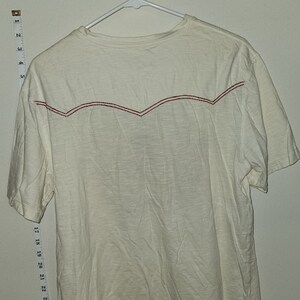 Puede incluir: Una camiseta blanca de manga corta con un dise&ntilde;o bordado rojo en la espalda. La camiseta est&aacute; colgada en una percha blanca.